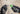A close-up of two gloved hands holding a Greenlee battery charger and a metal screwdriver against a blurred concrete background.