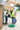 A construction worker wearing a white hard hat, safety vest, and knee pads kneels on a concrete floor, handling a green cable next to an electrical panel in a workshop setting.